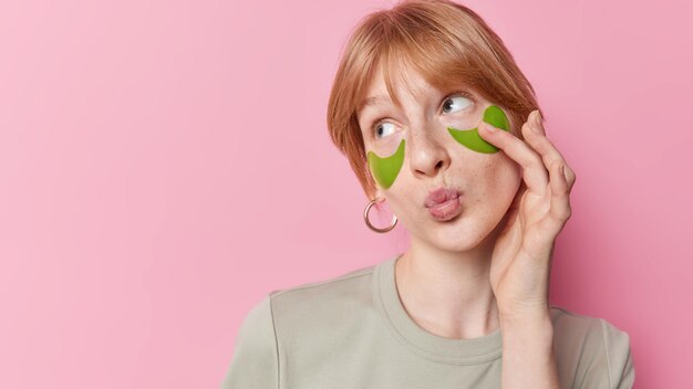 Photo of ginger girl with freckled skin keeps lips folded applies green hydrogel patches under eyes for skin treatment dressed casually isolated over pink background blank space for your promo