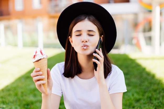 Photo of girl, talking on the cellphone, holding ice-cream in white t-shirt and hat on the street