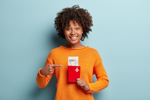 Photo of glad dark skinned curly female traveller points at tickets and passport, rejoices having trip abroad on summer holidays