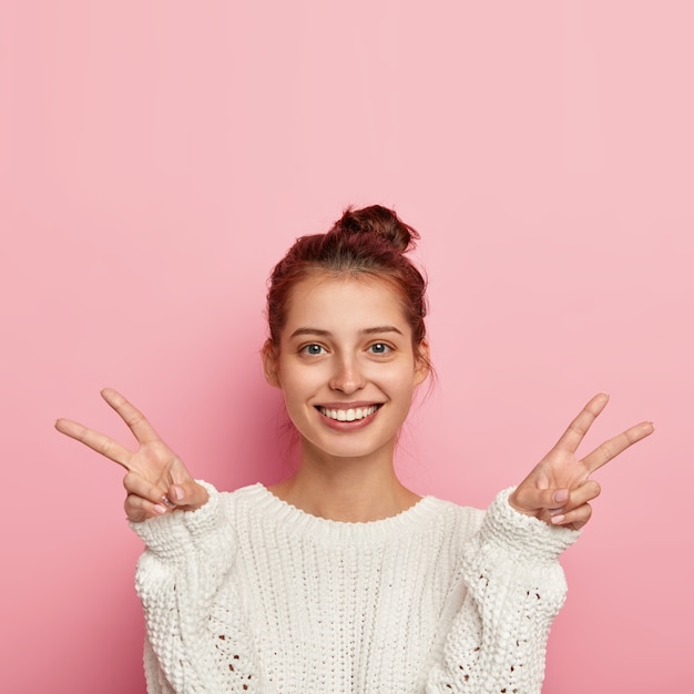 Foto gratuita la foto della donna europea felice sorride ampiamente, fa il gesto di vittoria della pace con entrambe le mani, ha un aspetto tenero, indossa un maglione bianco lavorato a maglia, esprime entusiasmo, mostra un segno di benessere, isolato sul rosa