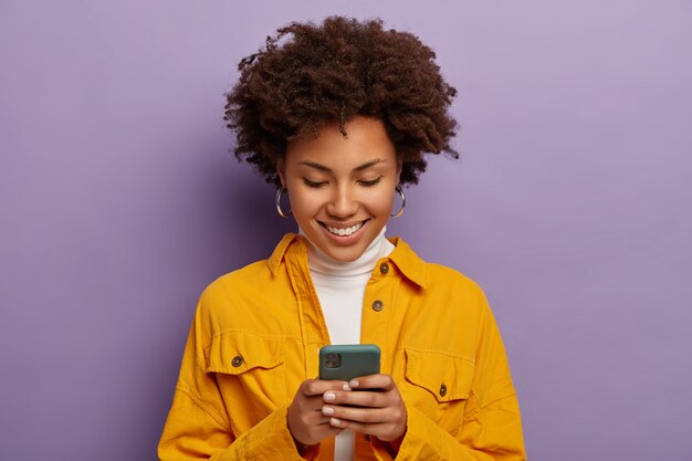 Photo of glad female, focused in smartphone, wears yellow shirt, smiles gently, isolated over purple studio wall.