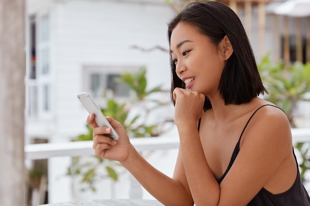 Photo of glad Japanese woman watches funny video on social networks via cell phone, sits in outdoor cafeteria, enjoys chat with boyfriend, texts message, uses free wifi, updates information.