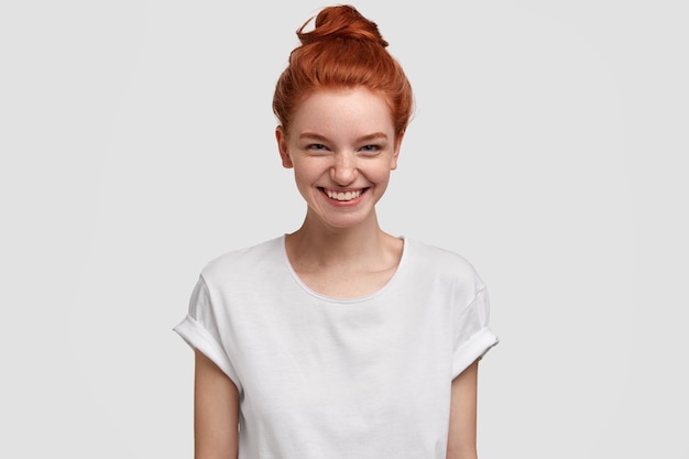 Photo of glad lovely red haired teenage girl smiles with curious and interested expression, accepts wonderful offer, wears casual white t shirt, models indoor