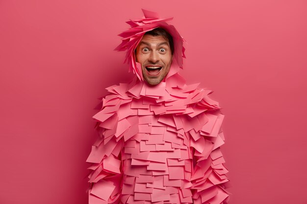 Photo of glad positive woman looks with wonder and happiness , has playful mood, giggles over funny joke, makes paper outfit of stickers, isolated on pink wall, has hilarious talk