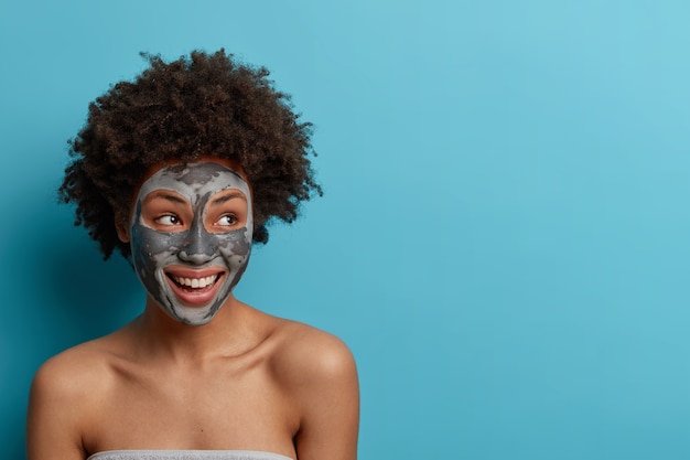 Photo of glad satisfied woman looks away with toothy smile, has cosmetic procedures at home, applies clay mask for skin care, shows bare shoulders, has well cared body. Blank space for your text