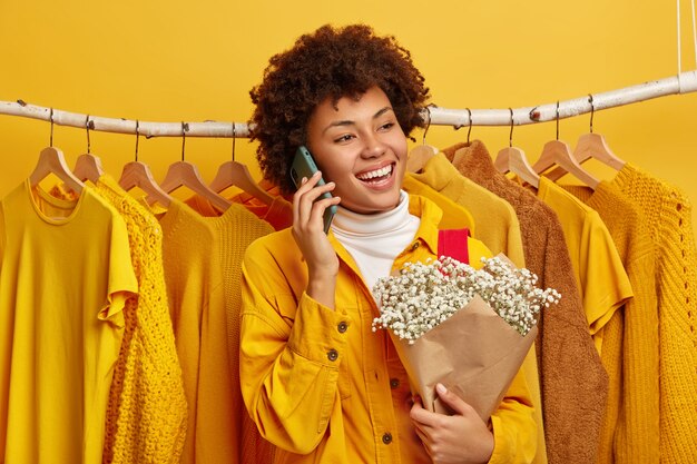 Photo of glad woman in bright yellow jacket, stands against clothes on hangers in her home wardrobe, ready for going out, calls friend via cellular holds bouquet. Female shopaholic likes yellow color