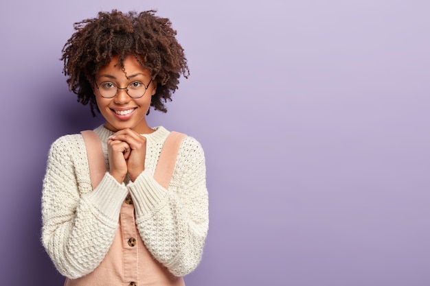 Photo of glad woman with dark skin, shows her teeth, keeps hands under chin, wears oversized white jumper, poses over violet  wall with blank space for your advertising content. emotions concept