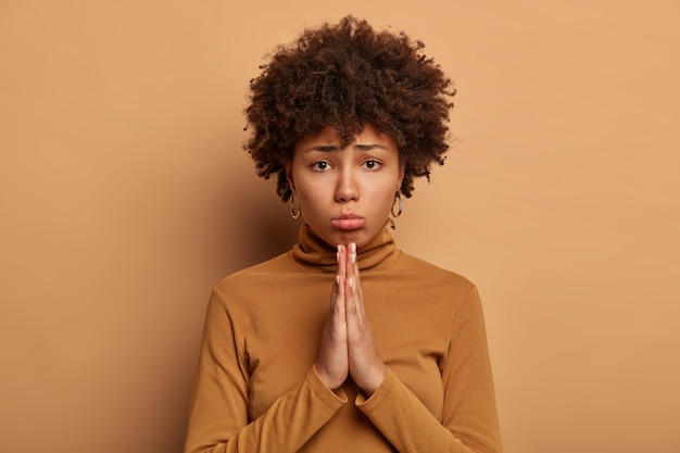 Photo of gloomy dark skinned curly woman asks for best, keeps palms pressed together, asks for forgiveness, dressed casually, isolated over brown wall, makes request. Please do me favor