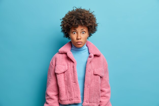 Photo of good looking adult woman has eyes popped out from wonder stares at camera hears shocking news wears blue turtleneck and pink coat poses indoor. Pretty ethnic girl feels confused shocked