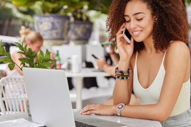 Photo of good looking African American female enterpreneur has work break in outdoor cafe, does remote work on laptop computer and makes call