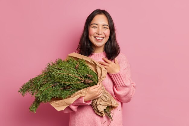 Photo of good looking brunette woman smiles broadly feels satisfied holds fir tree branches has festive mood going to make christmas composition wears winter jumper 