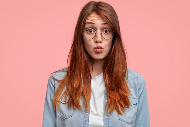 Photo of good looking female teenager with freckled skin, keeps lips round, makes grimace at camera, wears denim shirt, stands alone against pink background.