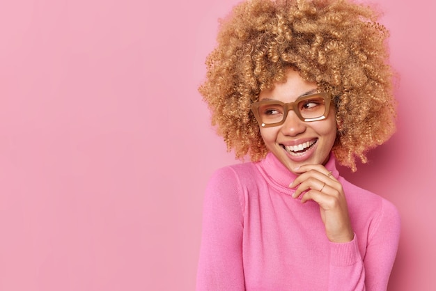 Foto di una bella donna tenera e sorridente che guarda felicemente lontano ha denti bianchi perfetti si sente felice indossa occhiali trasparenti e dolcevita isolato su sfondo rosa con spazio copia lontano