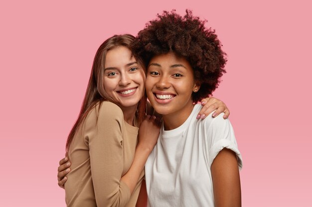 Photo of good looking two young models stand closely, embrace and enjoy togetherness, dressed in casual clothes, model over pink space