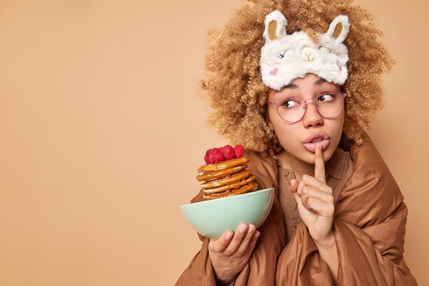 Photo of good looking woman makes silence gesture looks with mysterious expression away wrapped in soft blanket holds bowl of tasty pancakes for breakfast isolated over beige background blank space
