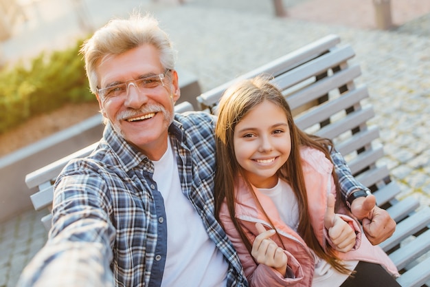 Photo of a grandfather and granddaughter sitting on a bench and make a selfie