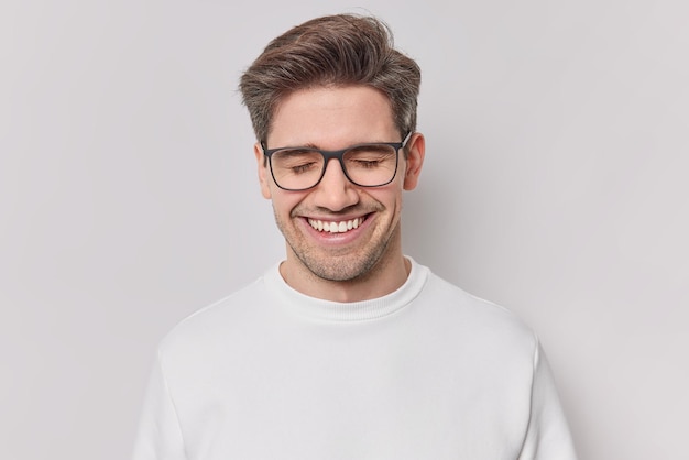 Фотография красивого жизнерадостного мужчины с темными волосами радостно улыбается с закрытыми глазами, выражает положительные эмоции, мечтает о чем-то, носит очки и повседневный джемпер, изолированный на белой стене