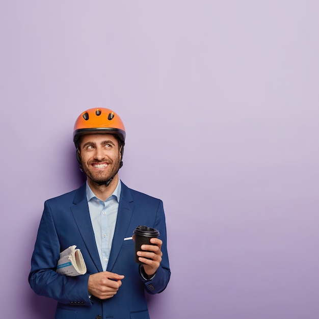 Photo of handsome cheerful man with toothy smile, carries crumpled newspaper, holds takeout coffee, thinks about new successful building project, wears protective helmet, formal suit