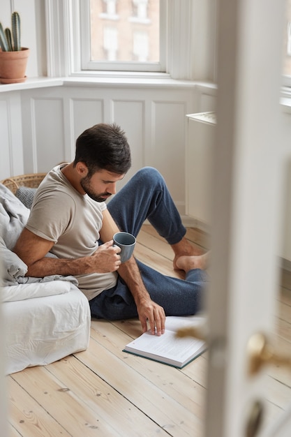 Photo of handsome guy relaxes with literature, enjoy relax at home, concentrated on reading, sits on floor, holds cup