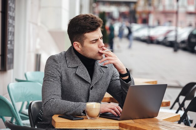 Photo of handsome man in gray coat smoking cigarette, and drinking cappuccino while resting in cafe outdoors
