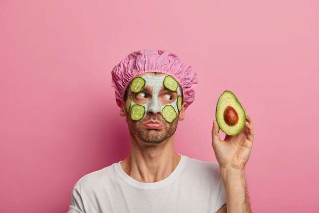 Photo of handsome man has rejuvenation facial treatment, holds avocado, applies fresh cucumber slices, wears bathcap