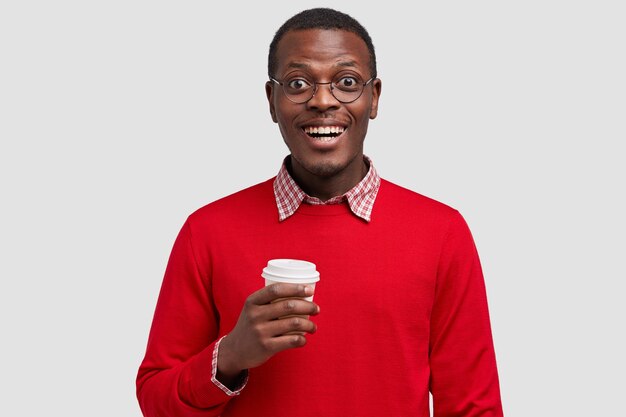 빨간 점퍼를 입은 잘 생긴 웃는 어두운 피부의 젊은 남자의 사진, 좋은 분위기에있는 테이크 아웃 커피를 보유하고 있습니다.