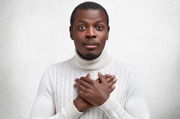 Photo of handsome young dark skinned male holds hands on chest as expresses gratitude, being pleased with heart piercing story