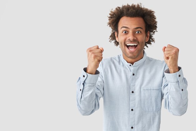 Photo of happy African American guy with positive expression, clenches fists with happiness, rejoices his success, dressed in elegant clothes, poses against white wall, copy space aside