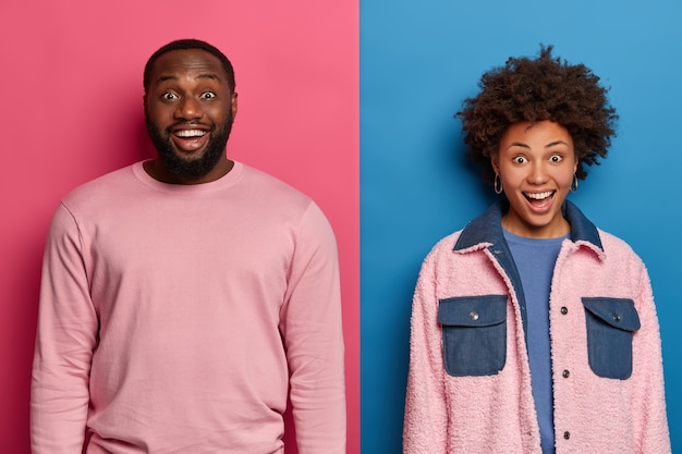 Фотография счастливой афро-американской пары стоят вплотную друг к другу, выражают положительные эмоции, радуются удивленному выражению лица, слышат отличные новости, вместе позируют на фоне розово-голубой стены.