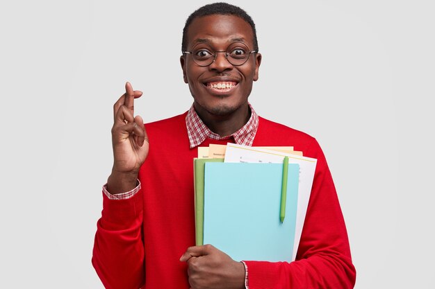 행복한 흑인 남자의 사진은 행운을 위해 손가락을 교차하고, 교과서를 들고, 붉은 옷을 입은 이빨 미소를 가지고 있습니다.