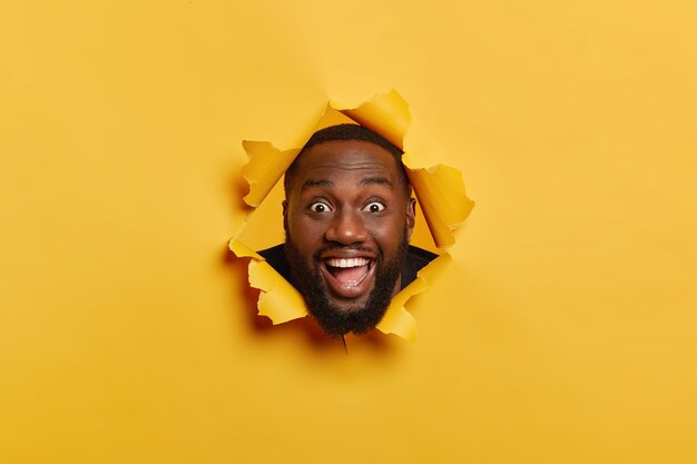 Photo of happy black man with pleased face expression, dark bristle, has fun indoor, keeps head in torn paper hole, laughs and looks at camera, isolated over yellow background.