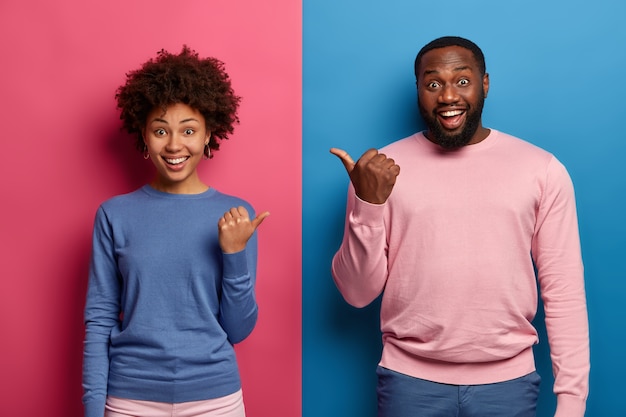 Foto gratuita la foto della donna di colore felice e del coniuge dell'uomo punta i pollici l'un l'altro, ha buon umore, suggerisce di sceglierne uno, sorride felice