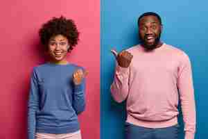 Free photo photo of happy black woman and man spouse point thumbs at each other, have good mood, suggests to choose one of them, smile happily