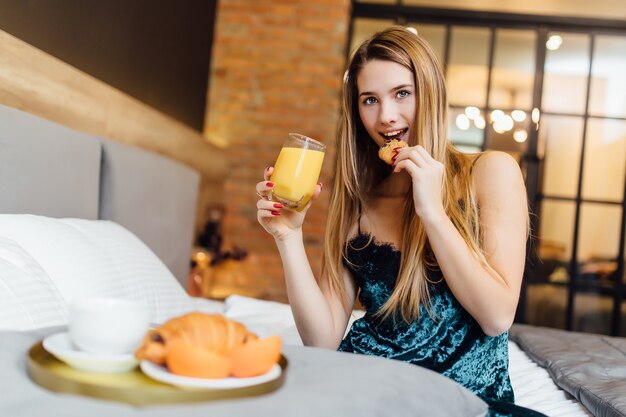幸せな金髪の女性の写真は、新鮮なオレンジジュースとクロワッサンと一緒に寝室で朝食をとります
