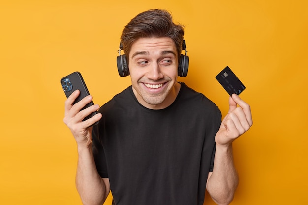 Photo of happy brunet man poses with credit card and smartphone orders in app pays online listens music via wireless headphones dressed in casuall black t shirt isolated over yellow background