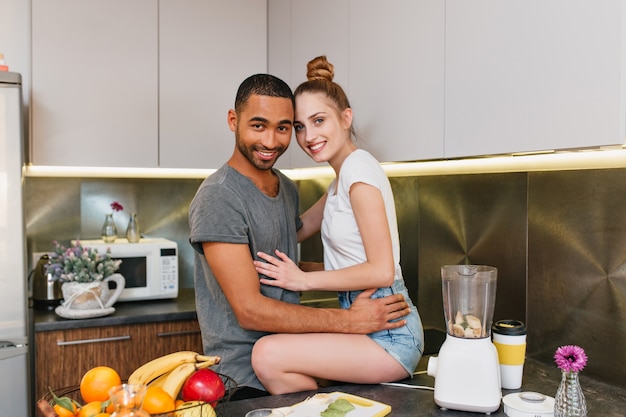 Photo of happy couple in the kitchen. Husband put his wife in shorts on the table. Lovers hugging each others. Sharing time at home, smile on faces.