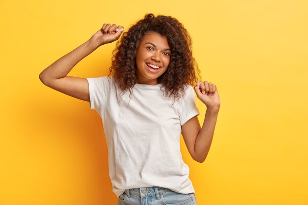 Photo of happy dark haired woman with positive expression, raises arms and moves while dancing, dressed in white casual t shirt and jeans