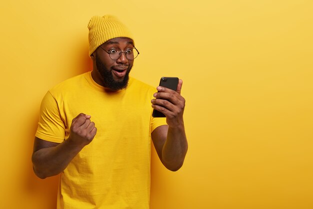 Photo of happy dark skinned guy celebrates victory of favourite team, reads results of game in internet, looks overjoyed at smartphone display