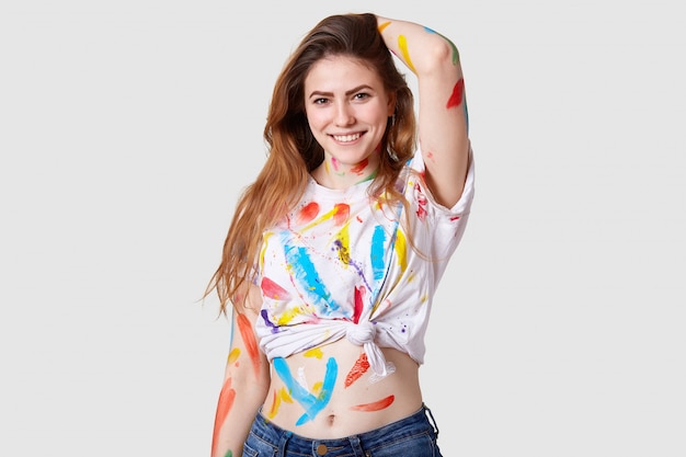 Photo of happy delighted female artist works on art ptoject, smiles positively, feels inspired and glad, has dirty stained top and hand with colourful paints, isolated over white  wall