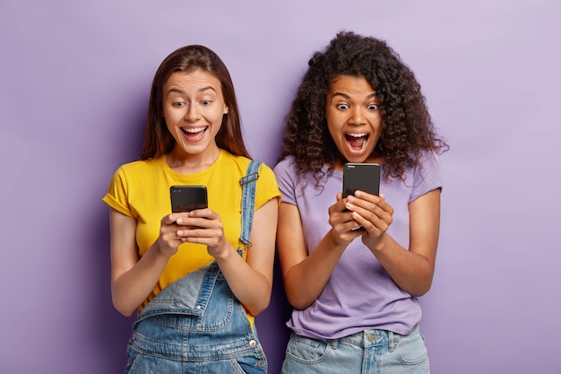 Photo of happy diverse female friends ignore live communication, chat in web blog via cell phones, gaze with positive expressions at screens