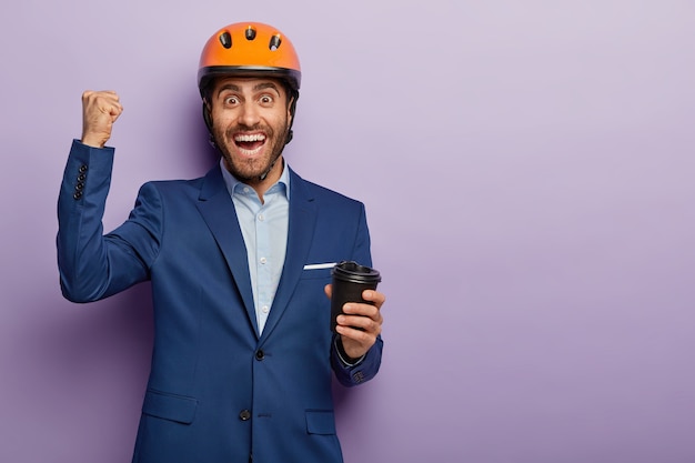 Photo of happy engineer has coffee break, holds paper cup, clenches fist with triumph, smiles gladfully, wears headgear and suit