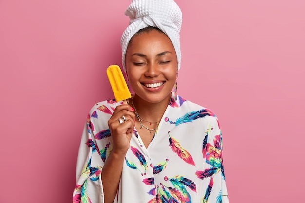 La foto della donna in buona salute felice con la pelle scura chiude gli occhi e sorride piacevolmente tiene il gelato delizioso