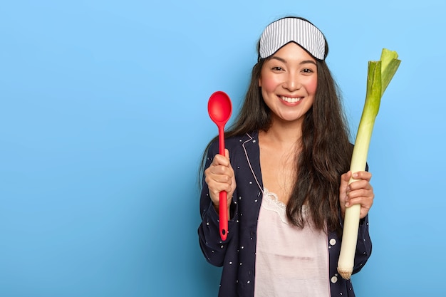 Photo of happy housewife going to cook healthy dish in morning, holds green leek and red spoon, smiles pleasantly, wears nightclothes