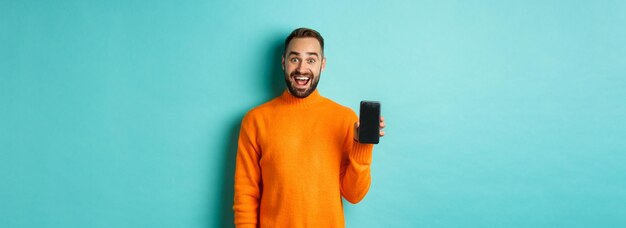 모바일 화면을 보여주는 행복한 남자의 사진은 청록색 위에 서 있는 온라인 상점 응용 프로그램을 소개합니다.