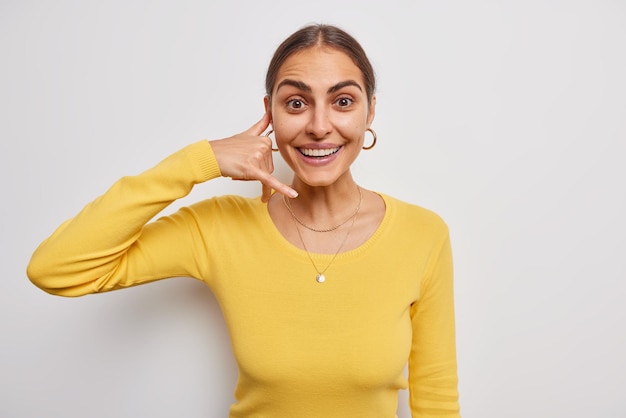 Photo of happy young European woman shows mobile phone call me gesture invites you and asks to contact her wears casual yellow jumper isolated over white wall