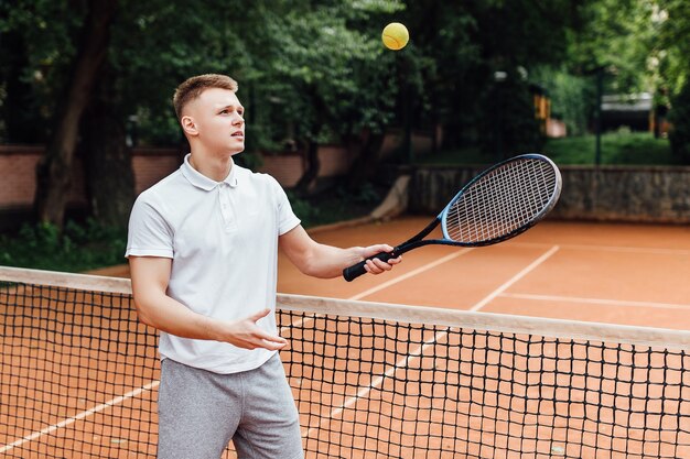 폴로 셔츠를 입은 행복한 청년이 테니스 라켓을 들고 테니스 코트에 서서 웃고 있는 사진.