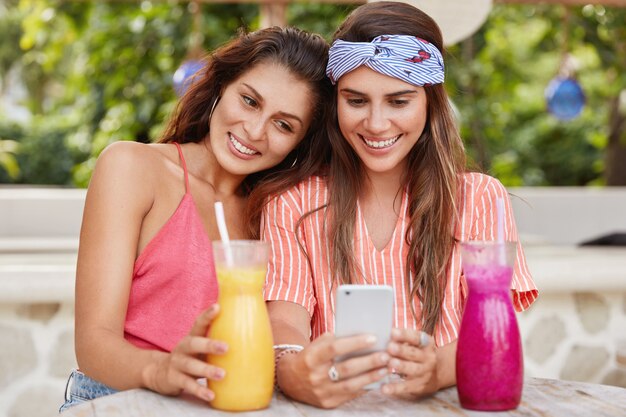 Фото счастливых молодых женщин, состоящих в однополых отношениях, просматривают интернет на мобильном телефоне, читают комментарии под постом, пьют свежие коктейли в кафетерии.