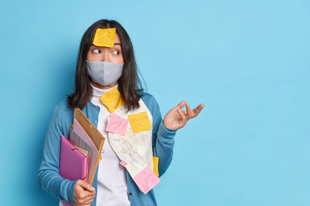 Photo of hesitant Asian woman shrugs shoulders doesnt know how to make reasearch work wears protective mask works during coronavirus pandemic.