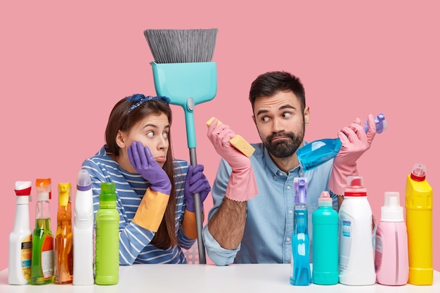 Photo of hesitant bearded man and displeased woman wears protective gloves, carries brush, work together, do household duties