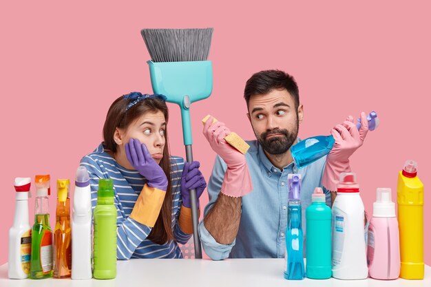 Photo of hesitant bearded man and displeased woman wears protective gloves, carries brush, work together, do household duties